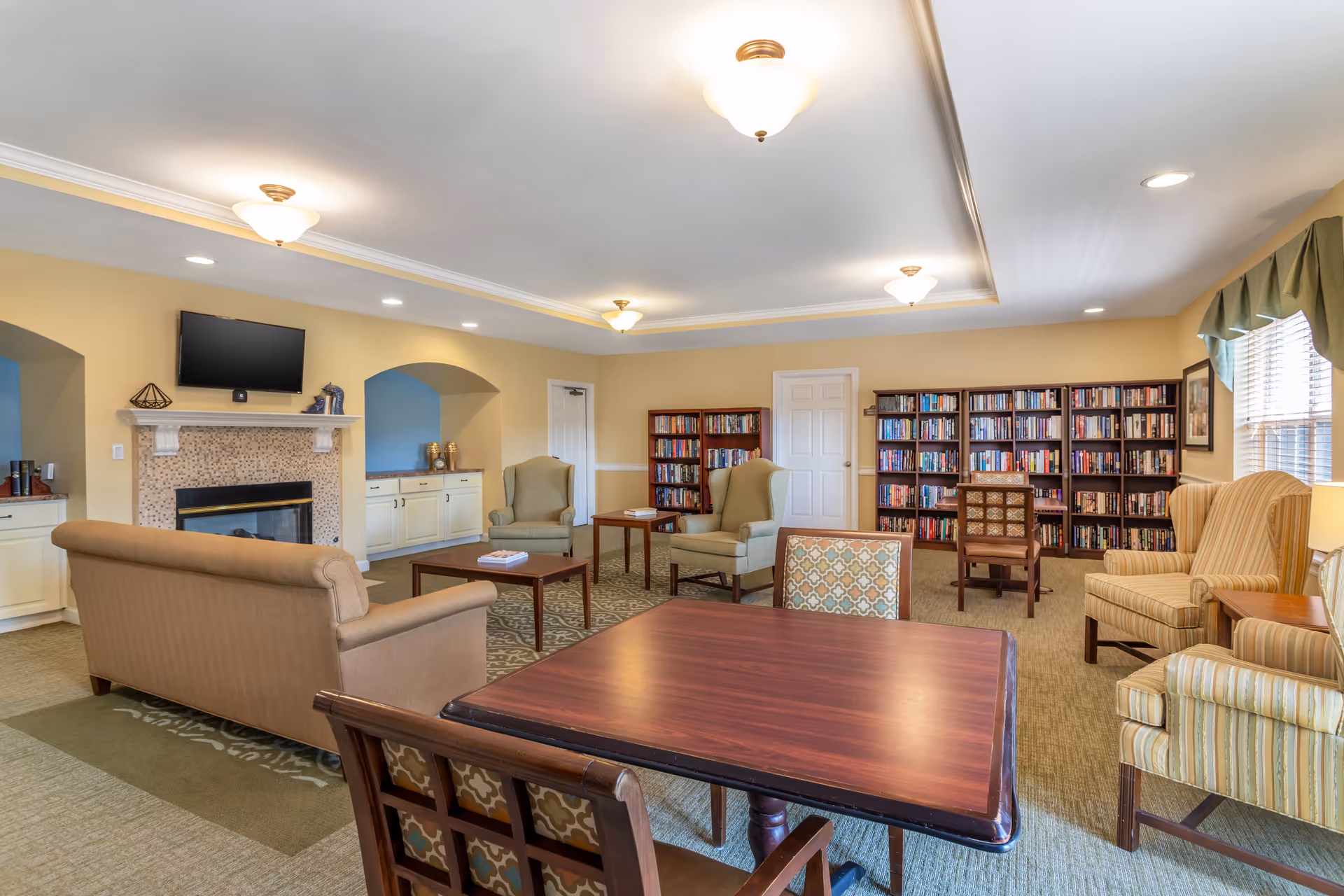 Bright senior living common room with sofas, armchairs, a fireplace, bookshelves and a central table.