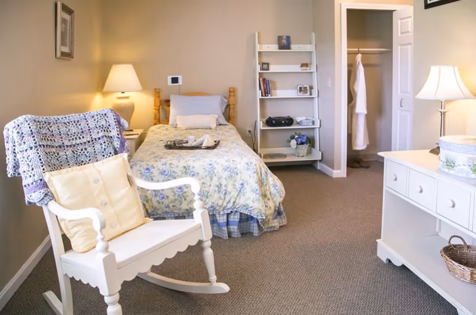 A cozy bedroom with a single bed covered in floral bedding, a white rocking chair with cushions and a crocheted blanket draped over it, a white dresser with a basket underneath, a white shelving unit with books and decor, and an open closet with a white robe hanging inside. The room is softly lit by two lamps.