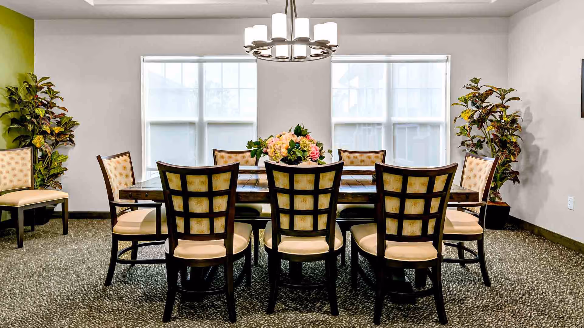 A dining room with a rectangular wooden table surrounded by eight cushioned chairs with wooden frames. The room has two large windows with white blinds, a modern chandelier hanging above the table, and two potted plants placed in the corners. A floral centerpiece is on the table.