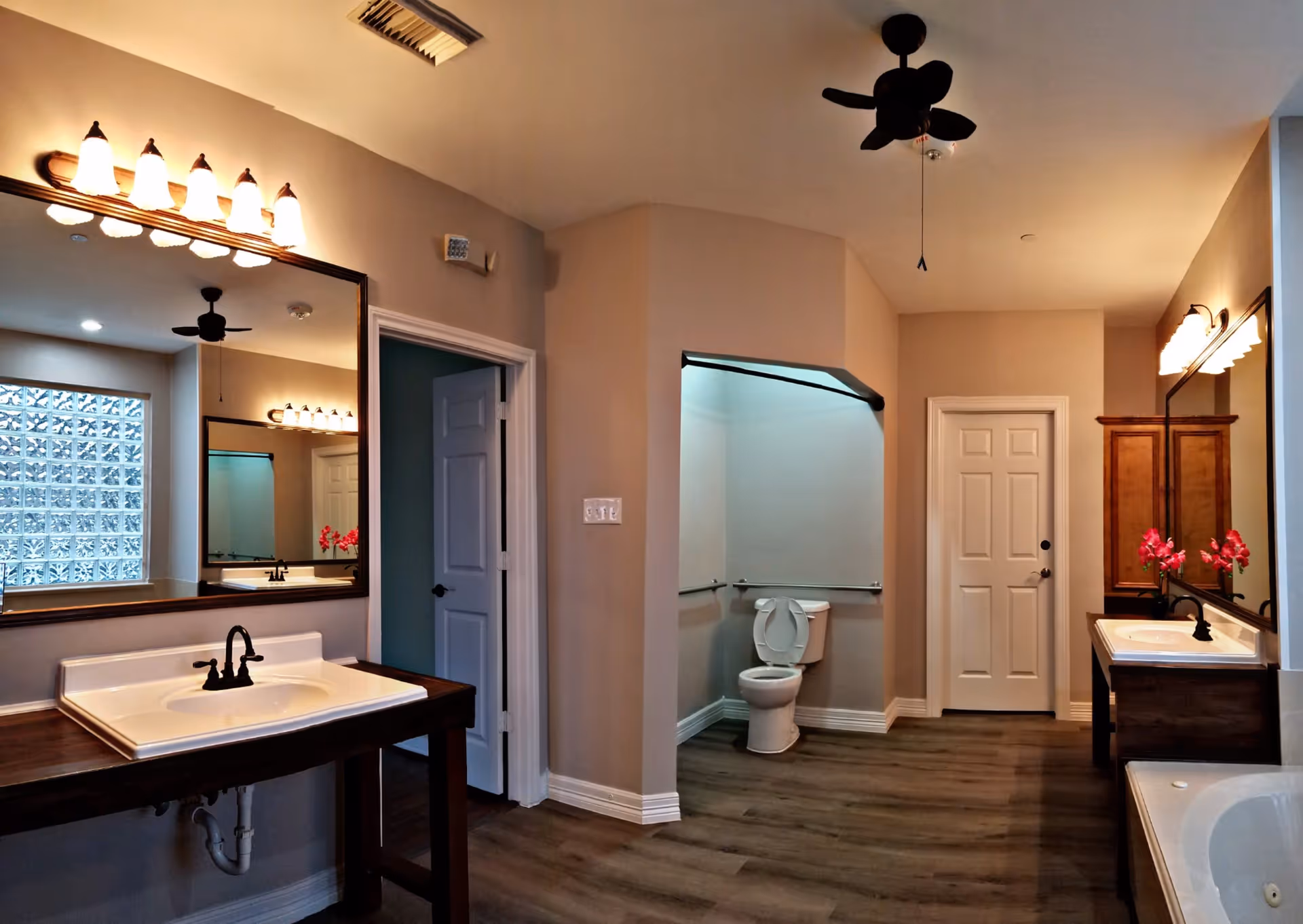 Spacious bathroom with double vanities, large mirrors, a toilet in an alcove, and a bathtub on wood-style flooring.