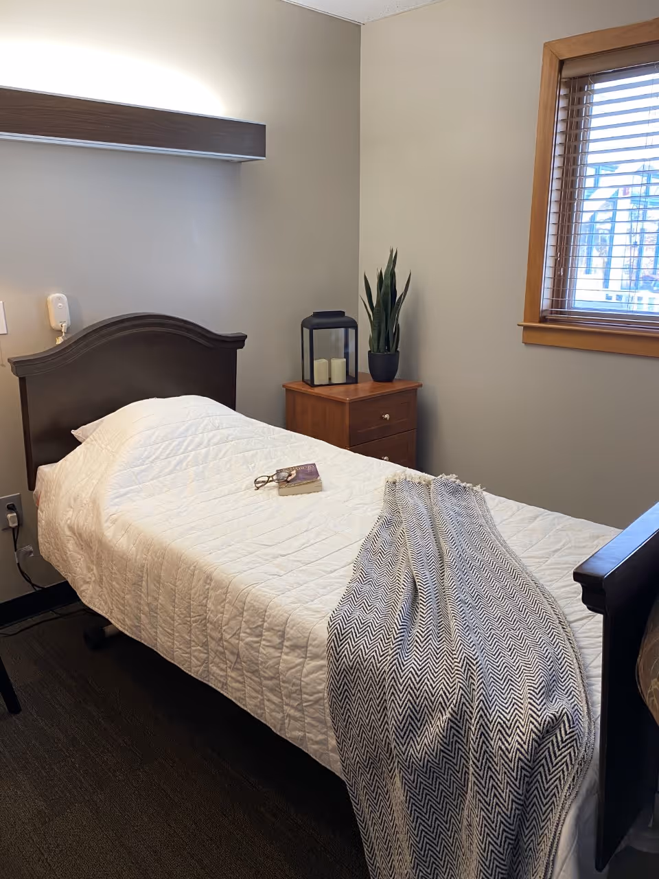 A small bedroom with a single bed covered in a white quilt. On the bed are a pair of glasses and a book, along with a folded patterned blanket at the foot. Next to the bed is a wooden nightstand with a black lantern containing three candles and a potted plant. A window with wooden blinds is on the right wall, letting in natural light.