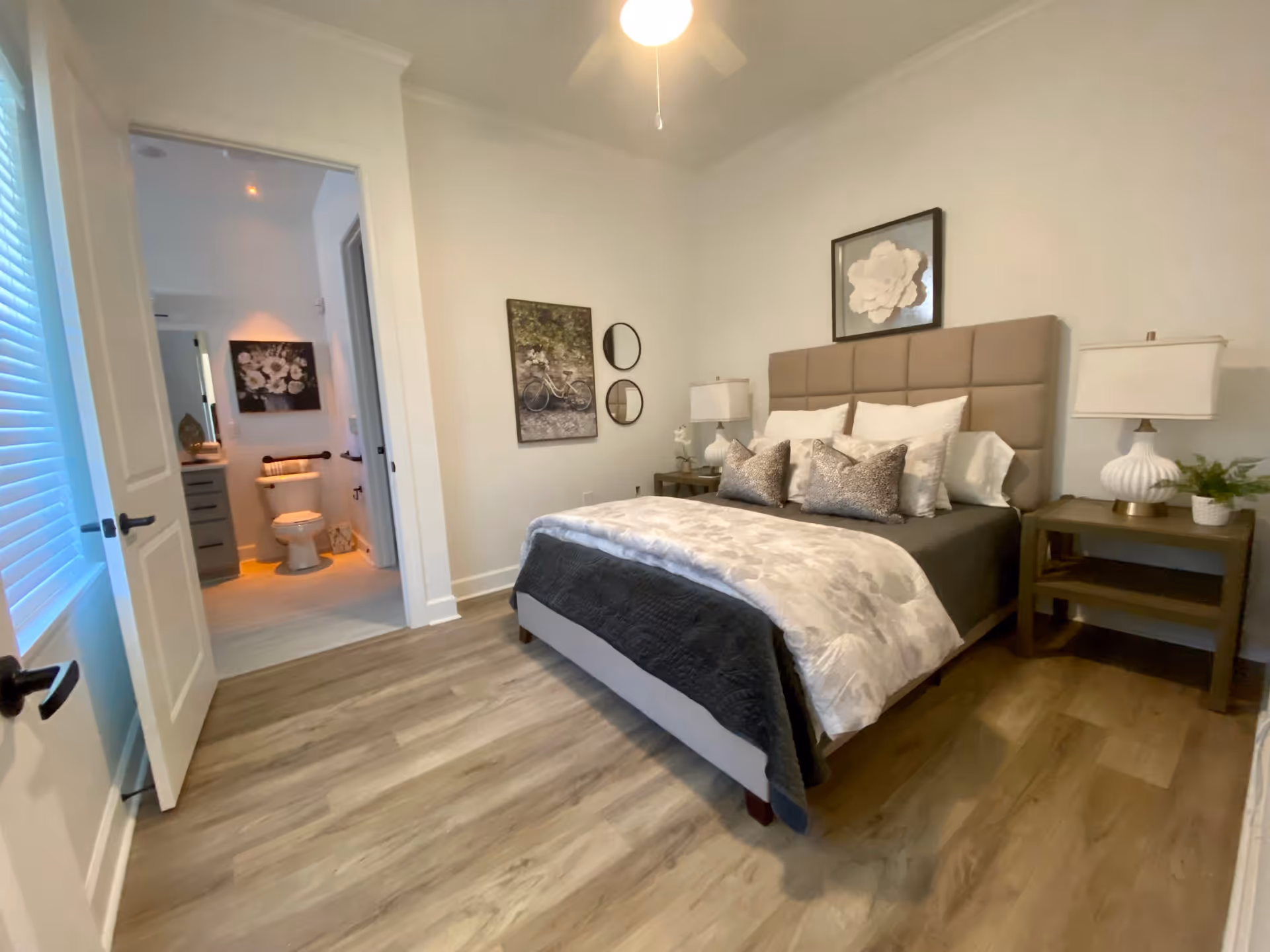 A neatly made bed with a padded headboard in a bedroom featuring two bedside tables with lamps and decorative items. The room has light-colored walls and wood flooring. An open door reveals a bathroom with a toilet, vanity, and wall art.