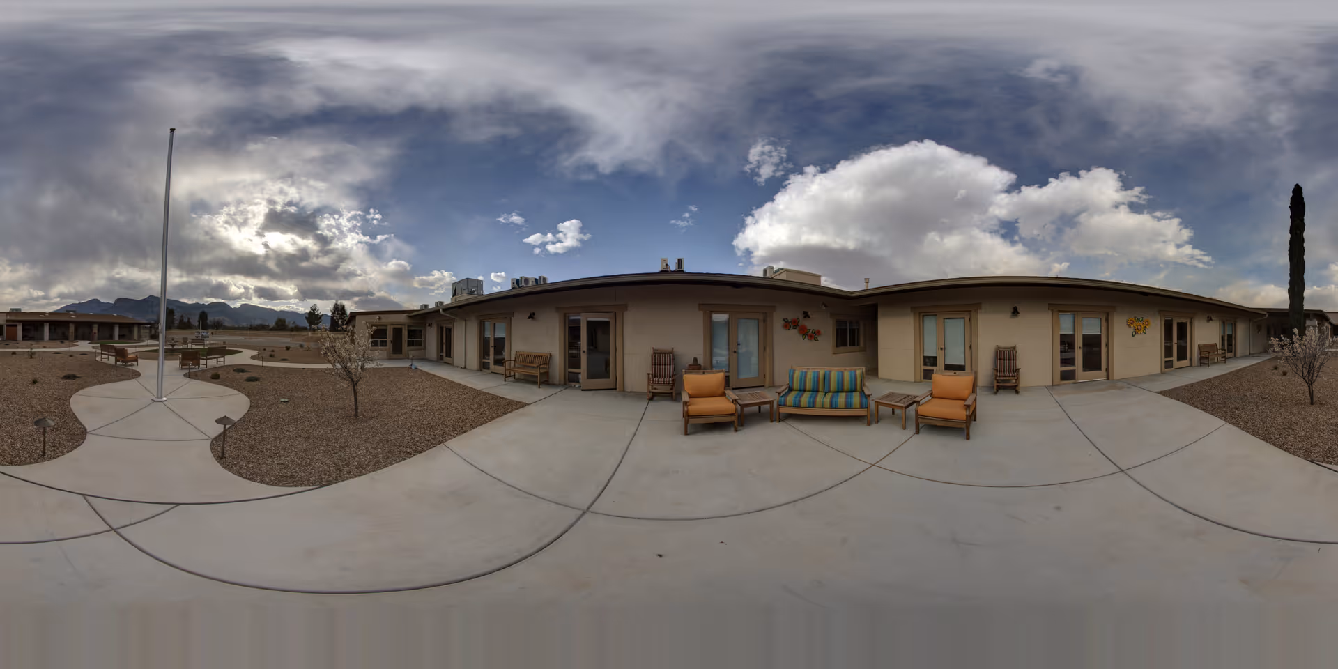 Outdoor courtyard area of Via Elegante Assisted Living Sierra Vista Canyons featuring a curved concrete walkway, several seating areas with chairs and a striped cushioned bench, small trees, and a flagpole under a partly cloudy sky with mountains in the background.