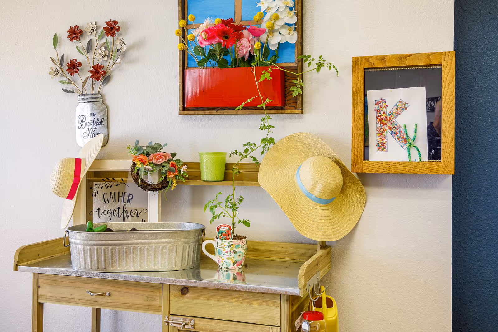 A decorative indoor gardening station with a wooden table holding a metal tub, a floral mug with a small plant, and a green pot. Two straw hats hang on the wall, along with colorful flower decorations and framed artwork, including a sign that says 'Gather together' and a letter 'K' made of colorful beads.