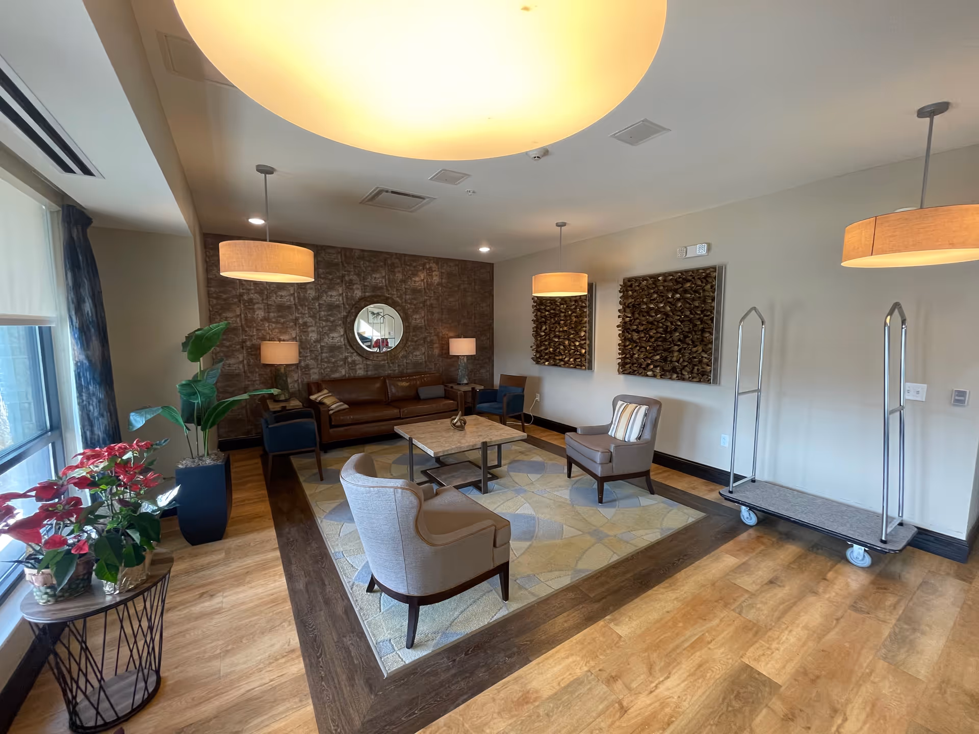 A well-lit lounge area with a brown leather sofa, two blue armchairs, and two beige armchairs arranged around a square coffee table on a patterned rug. The room features wooden flooring with a darker wood border, two large pendant lights, two table lamps, decorative wall art, a round mirror, and potted plants near a large window with curtains. A luggage cart is positioned against the wall.