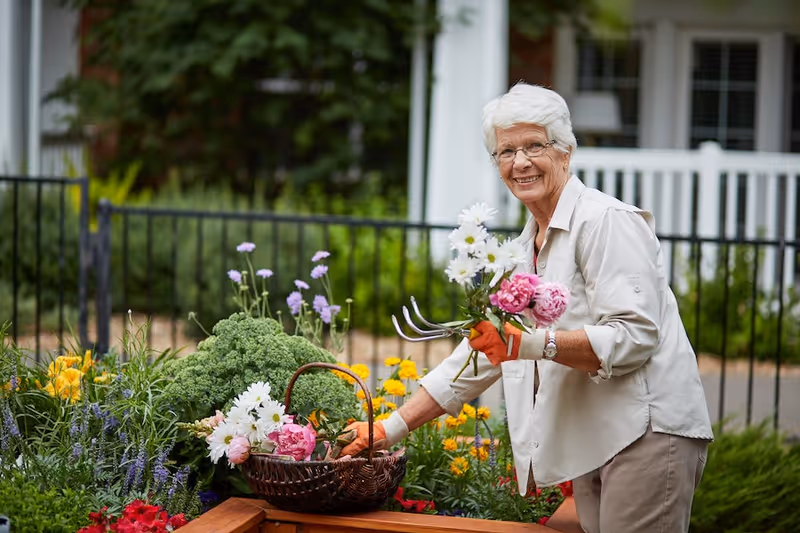 An elderly woman with white hair and glasses is gardening outdoors, holding a small bouquet of flowers including white daisies and pink peonies. She is wearing orange gardening gloves and a light-colored shirt, smiling while tending to a raised garden bed filled with various colorful flowers and greenery. A black metal fence and a white porch railing are visible in the background.