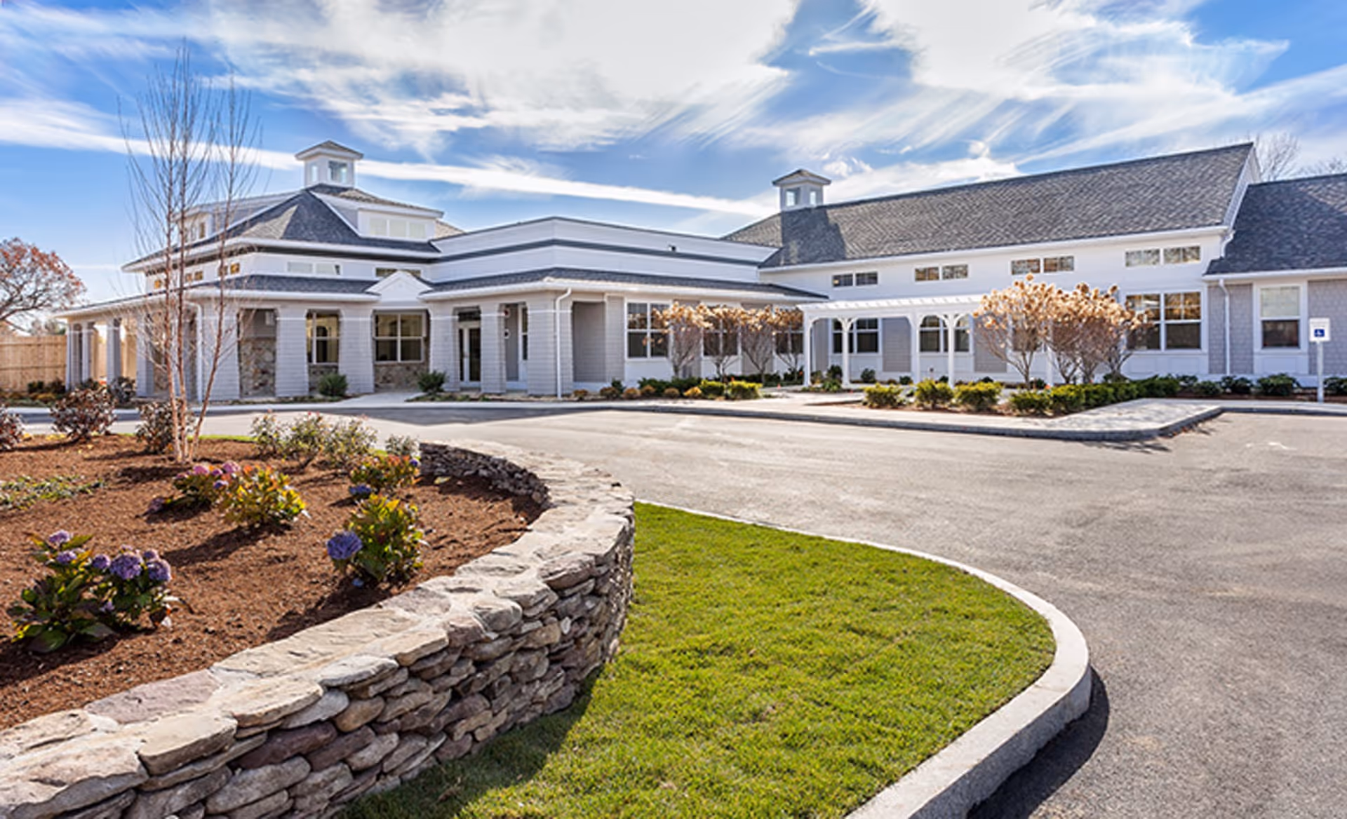 Exterior view of a single-story senior living facility building with a stone retaining wall and landscaped garden in the foreground under a partly cloudy sky.