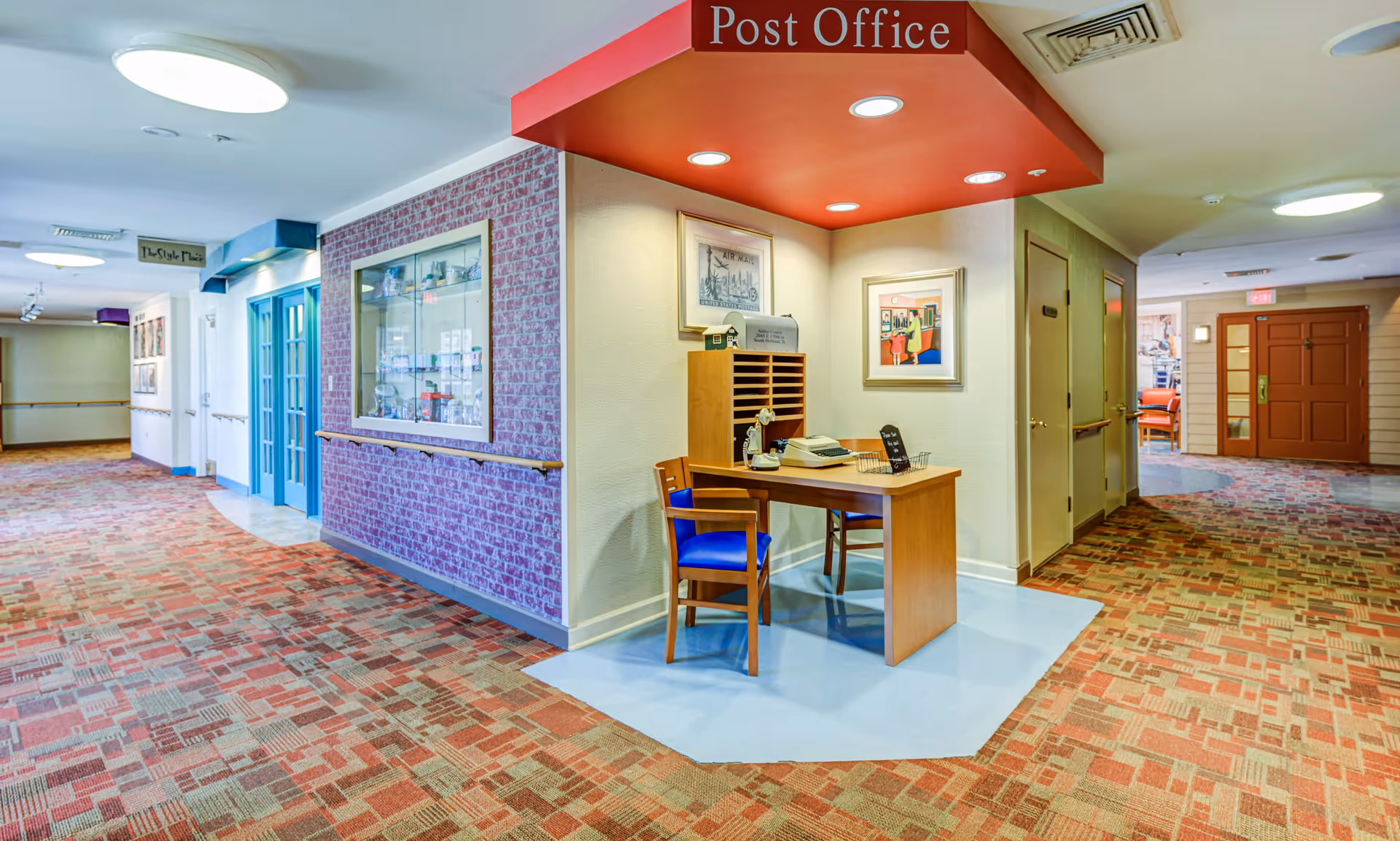 Interior hallway of a senior living facility with patterned carpet and a small post office area featuring a wooden desk, chair with blue cushion, mail slots, and framed artwork on the walls. The hallway has handrails and multiple doors leading to other rooms.
