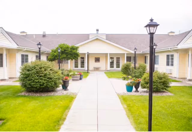 Outdoor courtyard area of Greene Senior Living facility with a paved walkway leading to a building entrance. The courtyard is landscaped with green grass, bushes, potted flowers, and several black lamp posts.