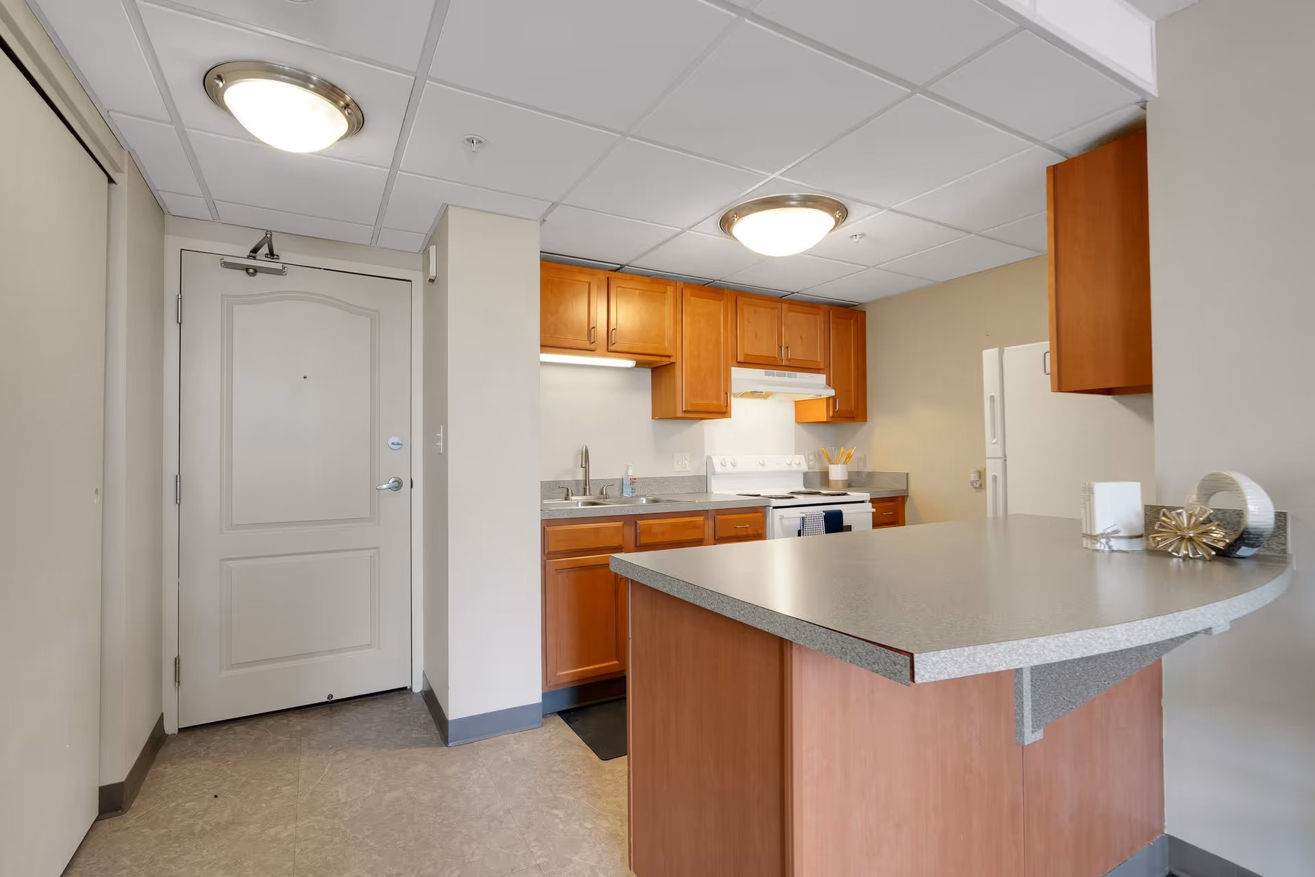 Interior view of a kitchen area in a senior living facility with wooden cabinets, a white stove and refrigerator, a sink, and a countertop extending into a breakfast bar. The entrance door is visible on the left side, and the ceiling has recessed lighting fixtures.