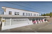 Exterior view of a single-story white building with a sign that reads 'Myrtle Point Care Center'. The building has several windows and a covered entrance area with pink flowers in planters. Power lines are visible above the building and there is a paved area in front.