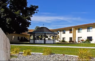 Outdoor view of a retirement living facility with a white gazebo in the center of a green lawn, surrounded by a two-story beige building under a blue sky.