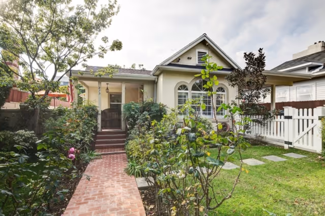 Front exterior of a single-story home with a brick walkway, small garden beds, a porch and white picket fence.