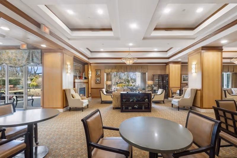 A spacious and well-lit common area in a senior living facility featuring multiple seating arrangements including round tables with chairs and comfortable armchairs. The room has a patterned carpet, wooden paneling, a fireplace, and large windows with a view of an outdoor garden area. The ceiling has recessed lighting and decorative beams.