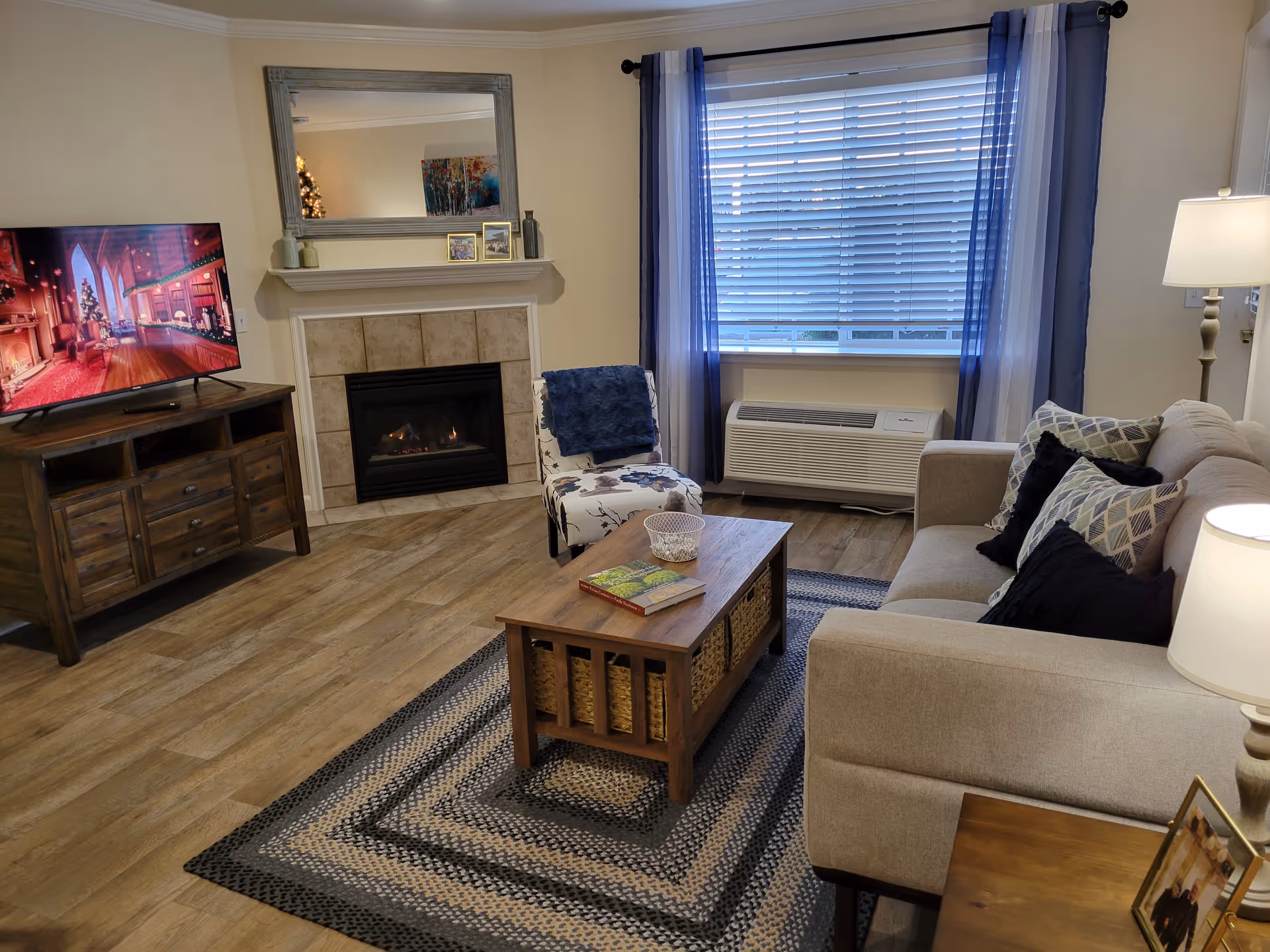 A cozy living room with a beige sofa adorned with patterned and solid pillows, a wooden coffee table with baskets underneath, a patterned armchair with a blue throw, a fireplace with a large mirror above it, a wooden TV stand with a flat-screen TV displaying a festive scene, a window with blue and white curtains, and a floor lamp beside the sofa.