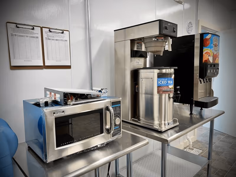 A small kitchen area with a stainless steel microwave, an iced tea dispenser, and a soda fountain machine on metal tables. Two clipboards with papers are hanging on the white wall in the background.