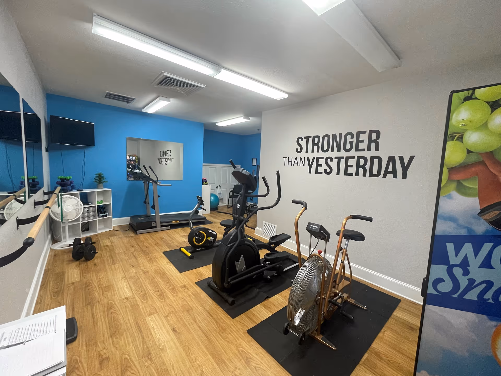 Small fitness room with cardio machines, free weights, mirrors, and a wall decal reading "Stronger Than Yesterday".