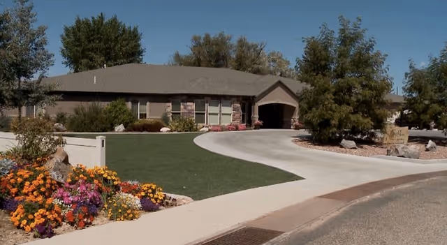 Single-story building with a sloped roof surrounded by a well-maintained lawn, colorful flower beds, and trees under a clear blue sky. A curved driveway leads to the entrance of the building.