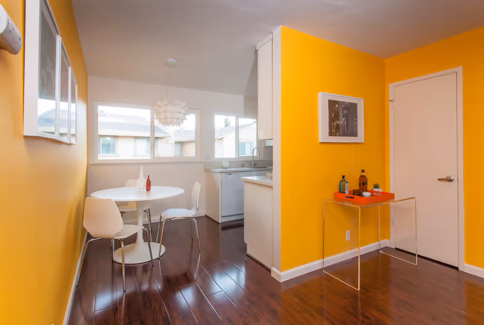 A bright dining area with yellow walls and dark wooden floors. There is a round white table with three white chairs around it, and two decorative vases on the table. The dining area is adjacent to a kitchen with white cabinets and a dishwasher. A small table with bottles and cups is placed against one yellow wall, above which hangs a framed black and white photograph. Large windows allow natural light to fill the space.