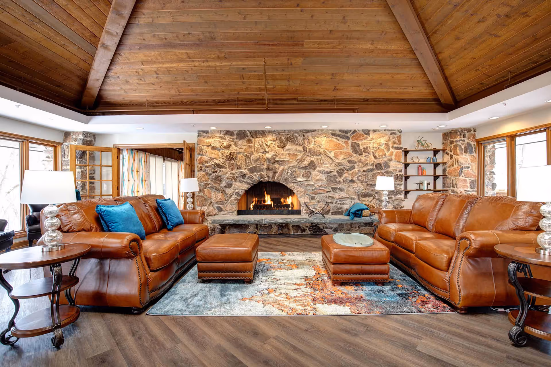 A cozy living room with two brown leather sofas facing each other, each adorned with blue throw pillows. Between the sofas are two matching leather ottomans on a colorful area rug. The room features a large stone fireplace with a fire burning, wooden ceiling beams, and large windows letting in natural light. There are side tables with lamps on either side of the sofas and a wooden shelf with decorative items on the right side of the fireplace.