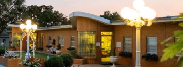 Single-story brick nursing home entrance at dusk with a curved roof, glowing lamp posts, and flowerbeds.