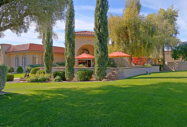 Exterior view of a senior living facility with a well-maintained green lawn, tall trees, and a building with a red-tiled roof and arched entrance. There are red umbrellas on a patio area in front of the building under a clear blue sky.