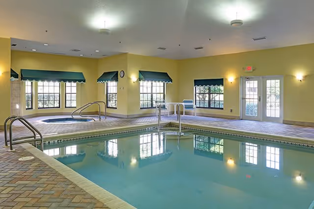 Indoor swimming pool area with a rectangular pool and a smaller circular hot tub in the corner. The room has yellow walls, multiple windows with green valances, tiled flooring, and ceiling lights. There are metal handrails for pool access and a door leading outside.