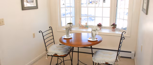 Small dining nook with a round wooden table and two cushioned metal chairs in front of a bright bay window.