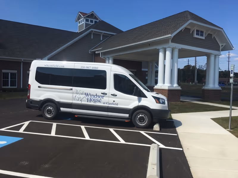 A white passenger van with the logo and text 'Windsor House at Canfield' parked in a designated parking area outside a building with a covered entrance supported by white columns under a clear blue sky.