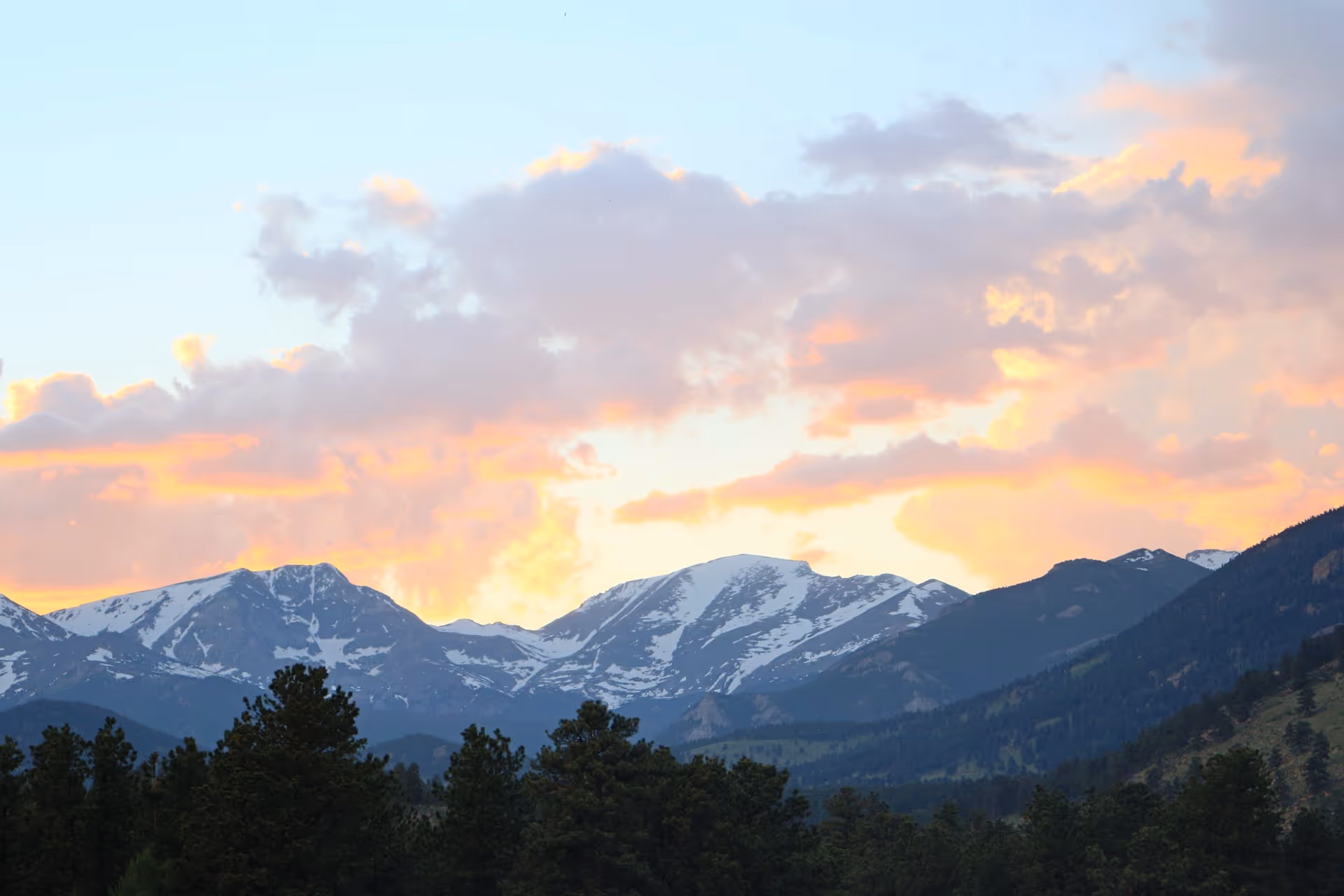 Scenic view of snow-capped mountains with a forest of pine trees in the foreground under a sky with orange and pink clouds at sunset.