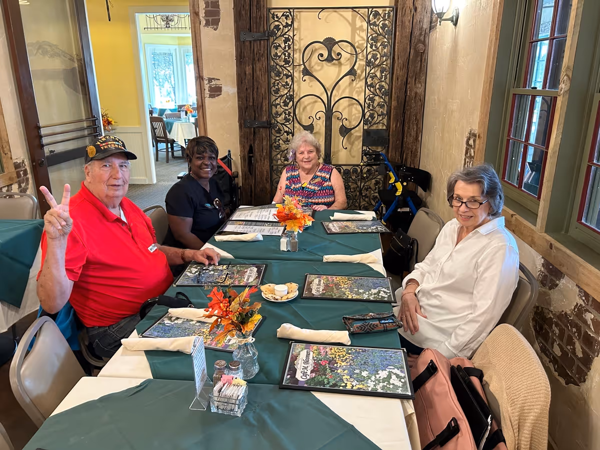 Four elderly people sitting around a table in a dining room with green tablecloths, menus, and small floral centerpieces. The room has rustic decor with exposed brick and wooden beams, and a decorative wrought iron gate on the wall behind them. One man in a red shirt and a veterans hat is making a peace sign.