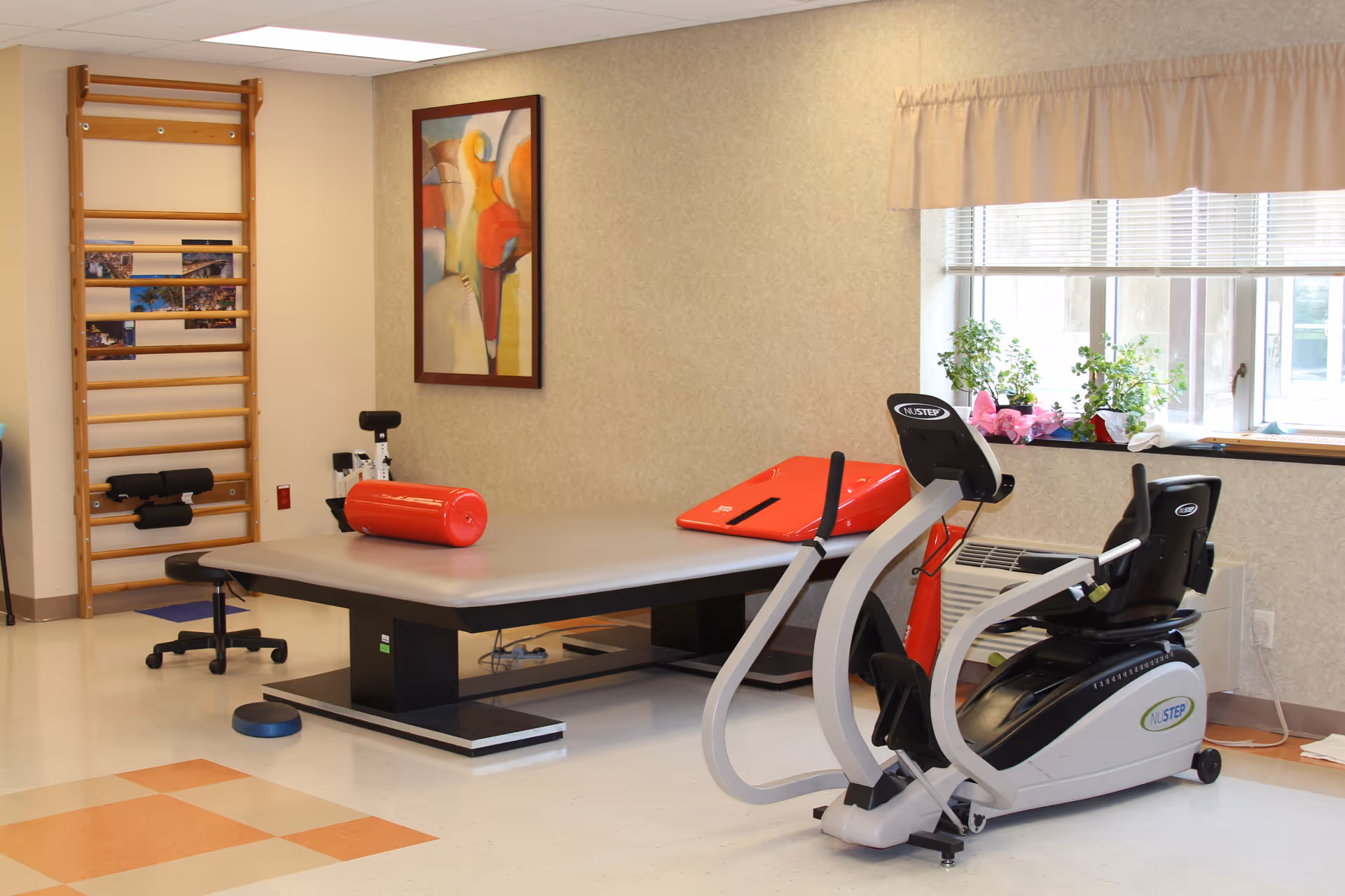 A physical therapy or rehabilitation room with exercise equipment including a NuStep recumbent cross trainer, a padded therapy table with red foam rollers, a wooden wall-mounted ladder, and a window with potted plants on the sill. The room has beige walls, a framed abstract painting, and a tiled floor with a checkered pattern.