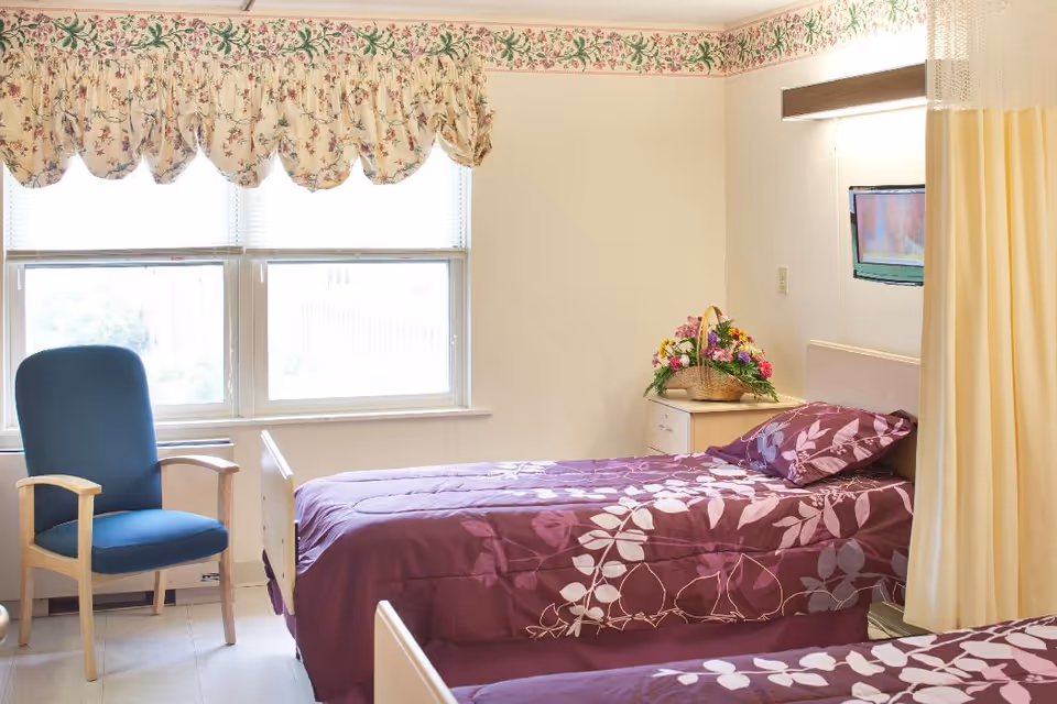 A cozy shared bedroom in a senior living facility featuring two beds, a chair, and decorative elements.