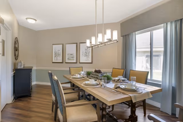 A dining room with a wooden table set for six people. The table has plates, cups, glasses, and napkins arranged neatly. There are six chairs around the table, and a modern chandelier hangs above it. The room has light-colored walls with three framed pictures and a large window with blue curtains letting in natural light. A small black cabinet is against one wall.