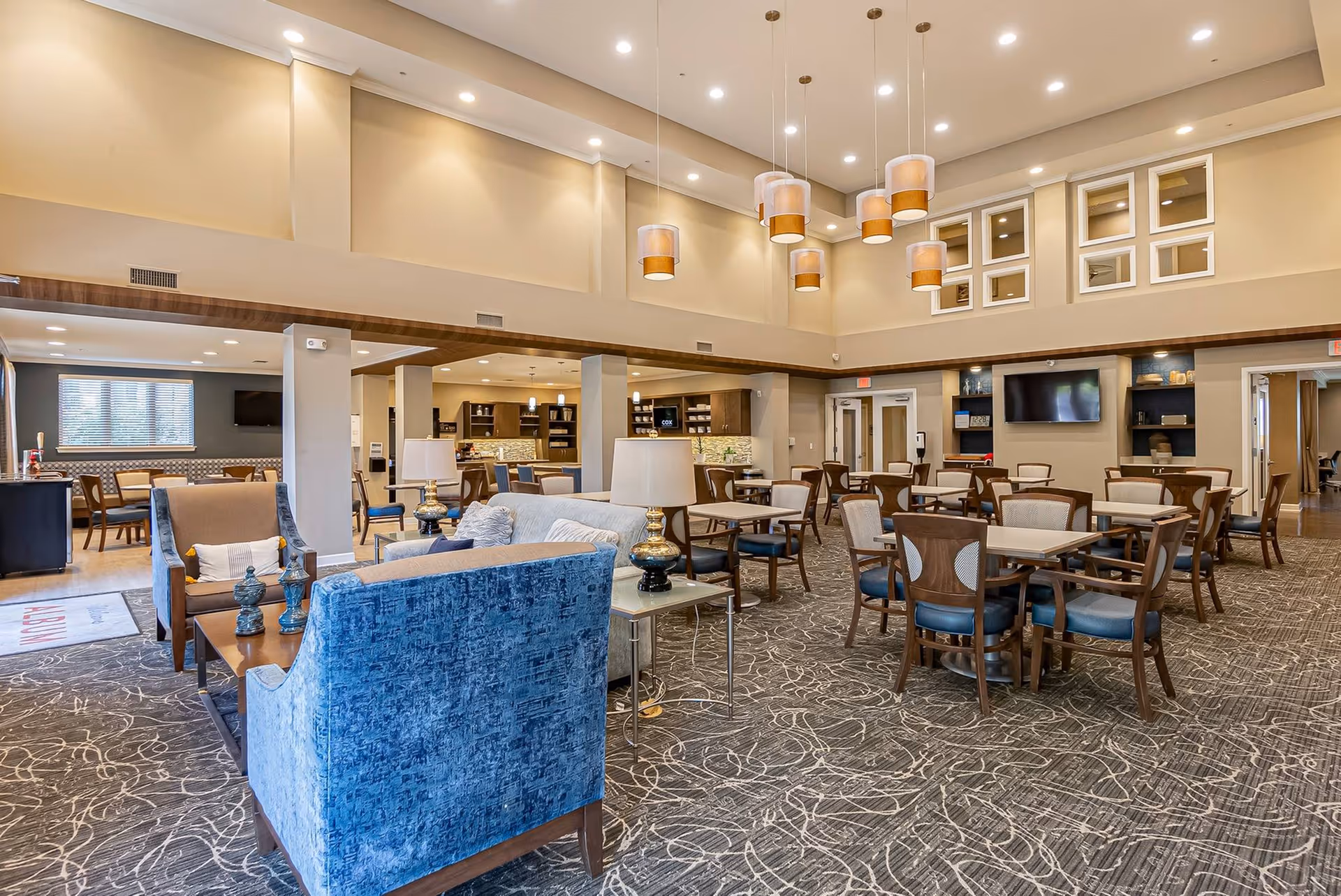 Spacious and well-lit common area in a senior living facility featuring multiple seating arrangements including blue and beige armchairs, wooden tables with chairs, decorative lamps, and modern pendant lights hanging from a high ceiling. The room has carpeted flooring with a patterned design, built-in shelves, and a television mounted on the wall.