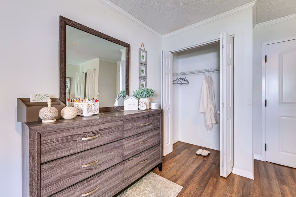 A bedroom interior featuring a wooden dresser with six drawers and a large mirror above it. On top of the dresser are decorative items including a small plant, an alarm clock, and containers holding pens and pencils. To the right is an open closet with a white robe hanging inside and a pair of slippers on the floor. The room has wooden flooring and white walls.