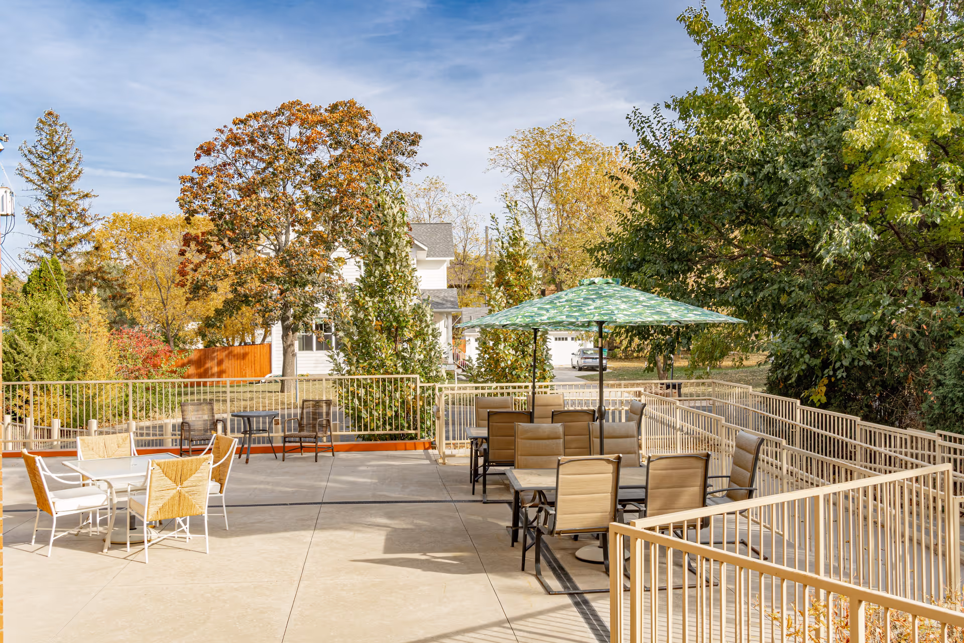 Outdoor patio area with multiple tables and chairs, including one table with a large green patterned umbrella. The patio is surrounded by beige metal railings and trees with autumn foliage in the background under a partly cloudy sky.