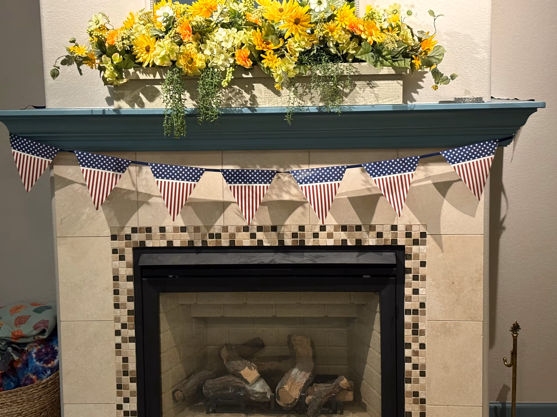 A decorative indoor fireplace with a tiled surround featuring small square mosaic tiles. The mantel above the fireplace is painted teal and adorned with a rectangular planter filled with yellow and white flowers and green foliage. Hanging below the mantel is a string of triangular American flag bunting. To the left, a basket with colorful blankets is partially visible.