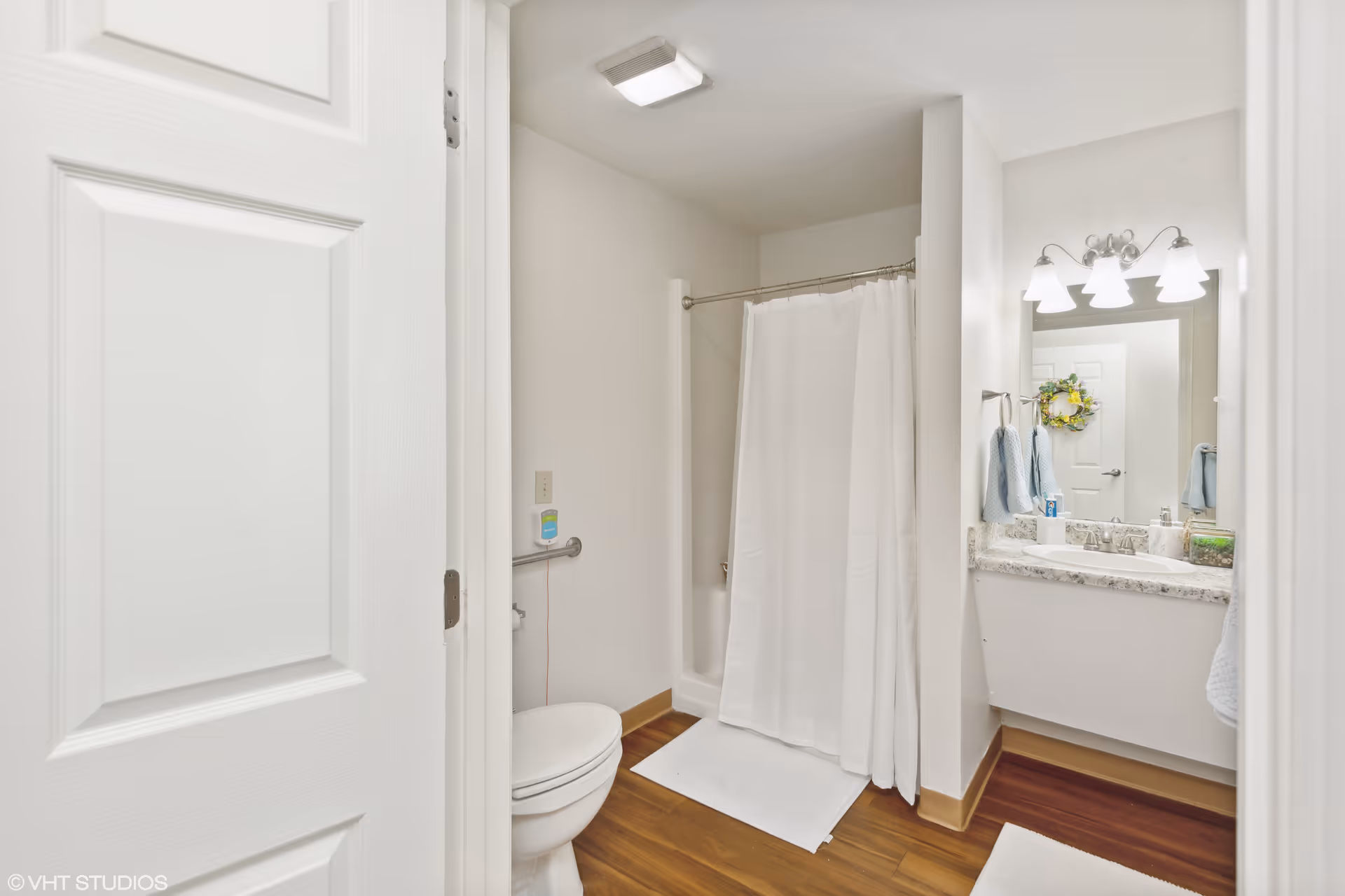 Small bright bathroom with a shower and curtain, toilet, and vanity with mirror and lights.