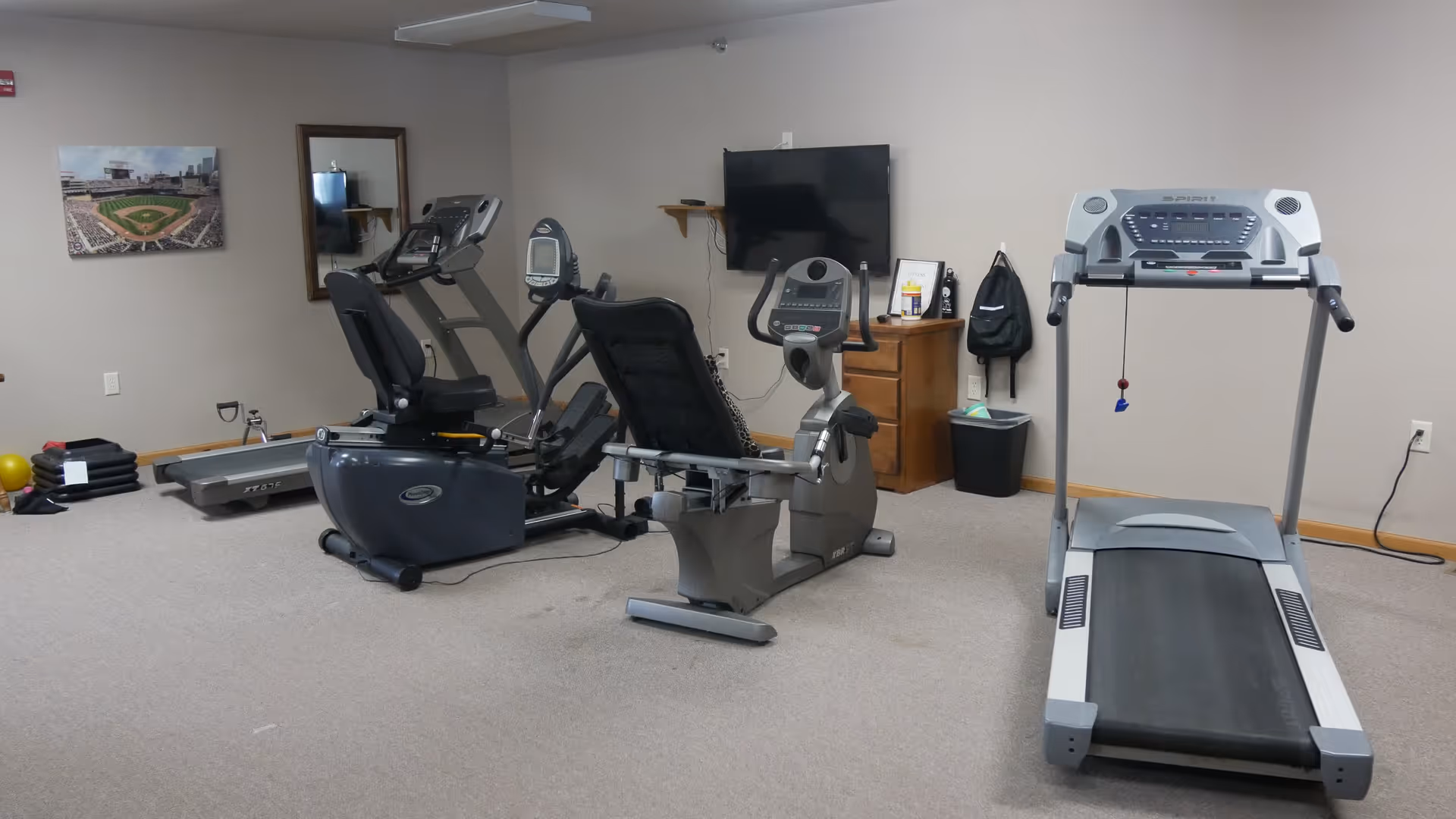 Indoor exercise room with a treadmill, two stationary bikes, and an elliptical machine. There is a wall-mounted TV, a wooden cabinet, a trash bin, and a backpack hanging on the wall. A framed picture of a baseball stadium is on the wall to the left.