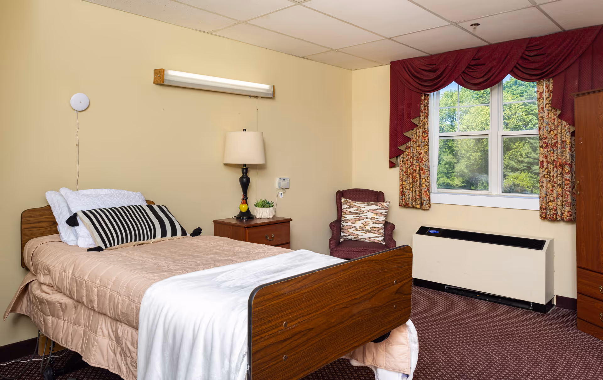 A single bed with beige bedding and a black and white striped pillow in a senior living facility room. Next to the bed is a wooden nightstand with a lamp and a small plant. There is a maroon armchair with a patterned cushion near a window with floral curtains and a maroon valance. Below the window is a heating or cooling unit. The walls are light yellow and the floor is carpeted in a dark red pattern.