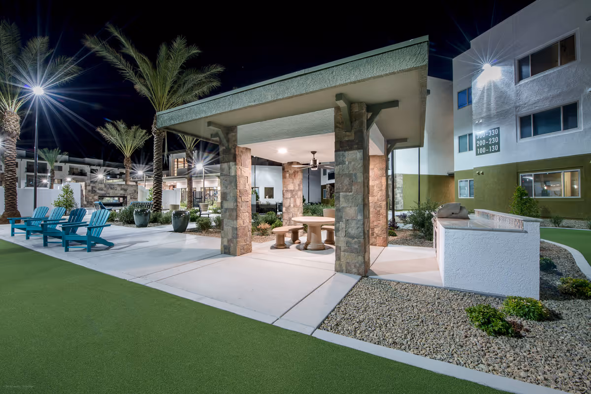 Night view of an outdoor seating area at a senior living facility with a covered pavilion supported by stone pillars, a round table with benches underneath, turquoise Adirondack chairs on a concrete patio, palm trees, and a barbecue grill. The building in the background has illuminated windows and signs indicating room numbers.