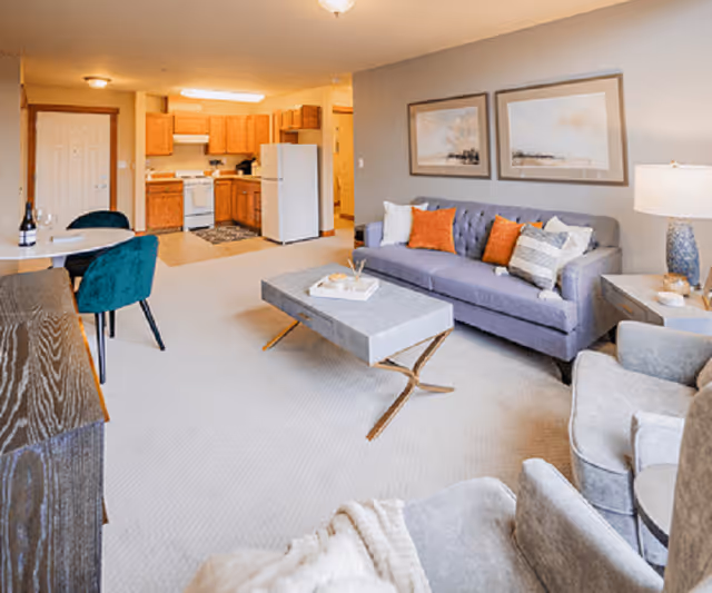 A bright and cozy living area in a senior living facility featuring a gray couch with orange and striped pillows, two gray armchairs, a modern coffee table, and a side table with a lamp. In the background, there is an open kitchen with wooden cabinets, a white refrigerator, stove, and a small dining table with two teal chairs.