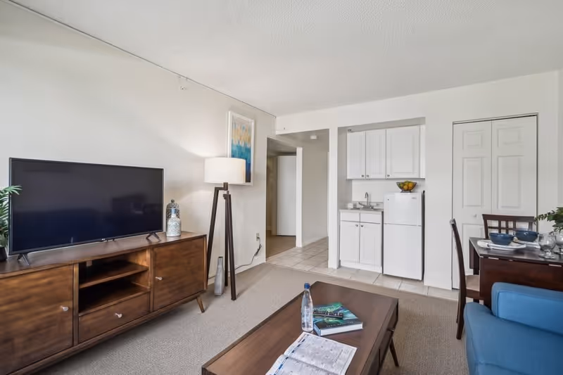 A cozy living room area with a flat-screen TV on a wooden stand, a floor lamp, a coffee table with books and a water bottle, a blue armchair, and a small kitchenette with white cabinets and a refrigerator. A dining table with chairs is partially visible on the right.