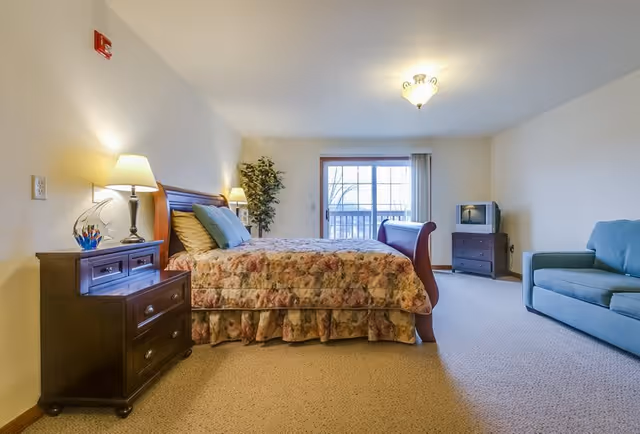 A spacious senior living bedroom with a large bed covered in a floral bedspread, two bedside tables with lamps, a potted plant in the corner, a sliding glass door leading to a balcony, a small TV on a dresser, and a blue couch.