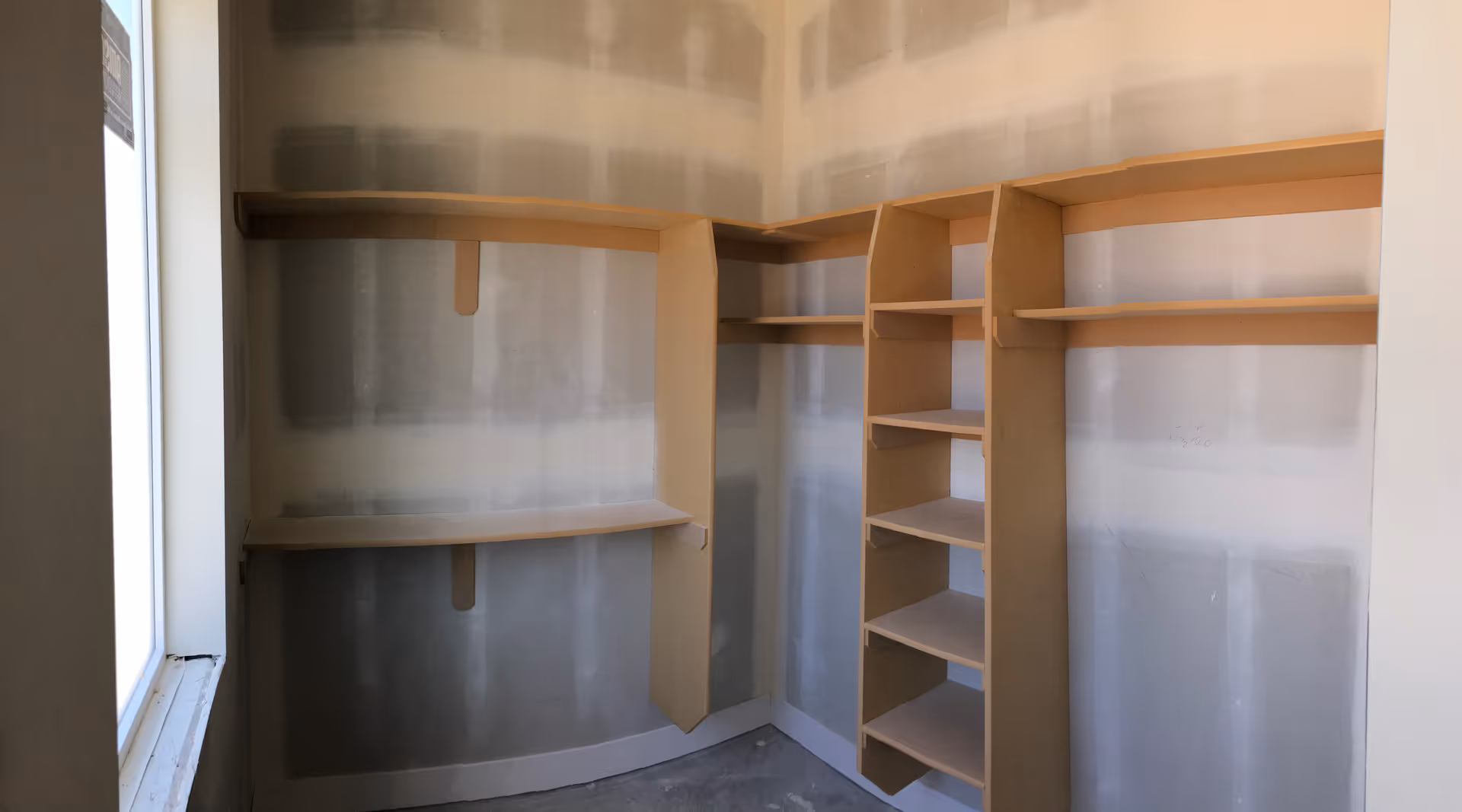 An empty walk-in closet with unfinished drywall walls and built-in wooden shelves and hanging rods. There is a window on the left side allowing natural light into the space.