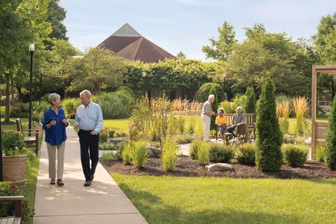 Two seniors walk along a landscaped path while others sit and talk on benches in a sunny garden courtyard.