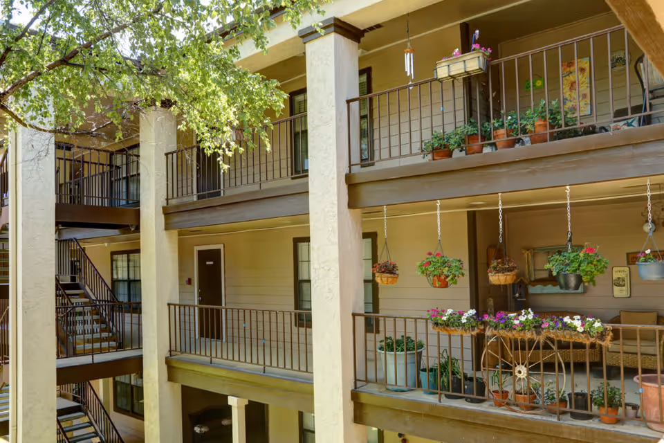 View of an apartment building's exterior hallway with two levels of balconies decorated with various potted plants and hanging flower baskets. There is a staircase on the left side and a tree with green leaves partially shading the area.