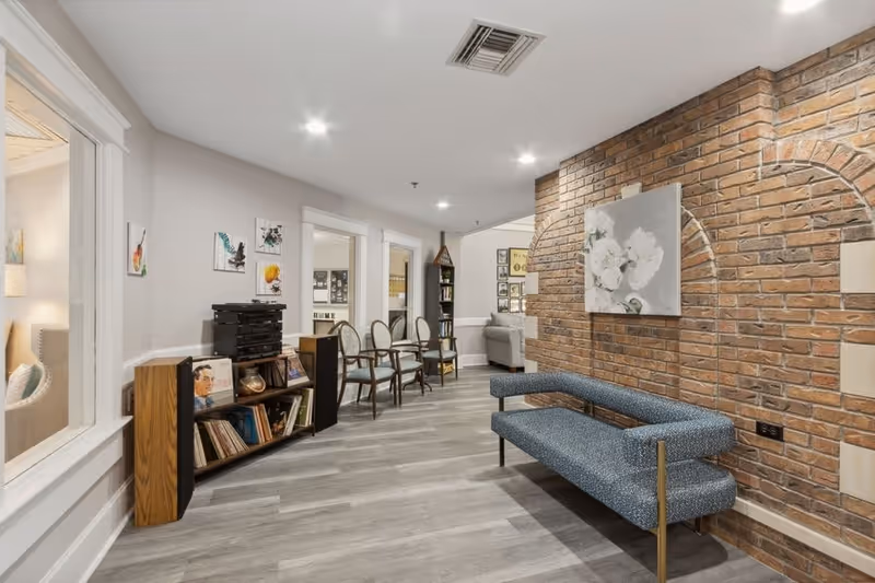 Interior view of a senior living facility hallway with a brick accent wall featuring a painting of white flowers. There is a modern blue bench along the brick wall and several wooden chairs with cushions lined up against the opposite wall. A bookshelf with records and a stereo system is also visible. The floor is light gray wood, and the ceiling has recessed lighting.