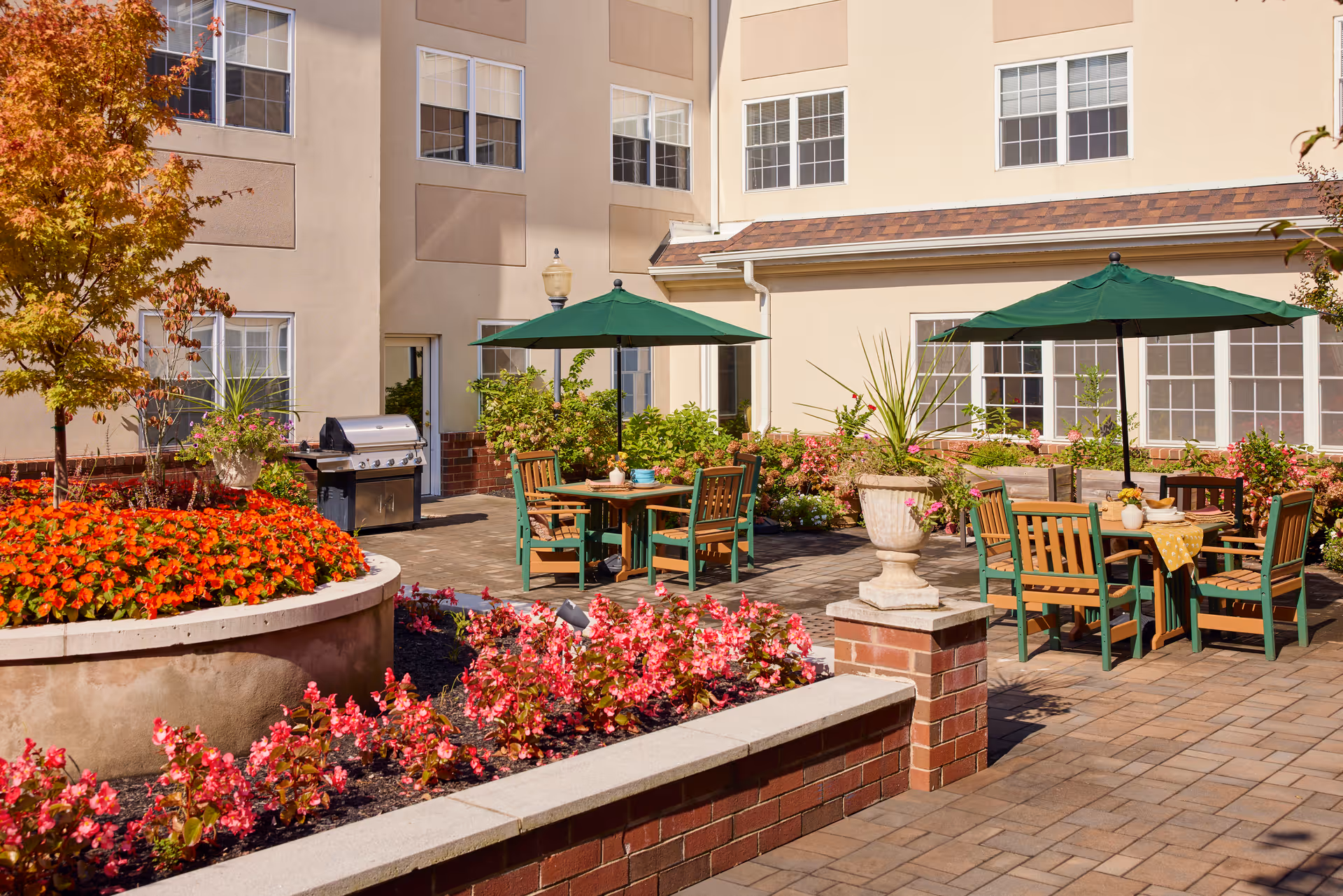 Outdoor patio area at Brandywine Mountain Ridge by Monarch with tables and chairs under green umbrellas, surrounded by vibrant flower beds and a barbecue grill near the building.